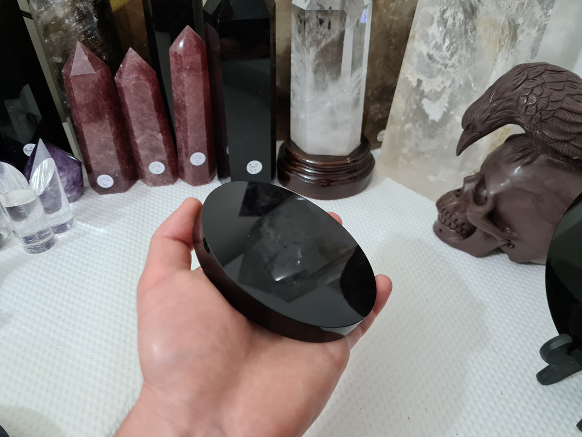 A hand holding a Black Obsidian crystal scrying mirror that is 10 centimetres in size